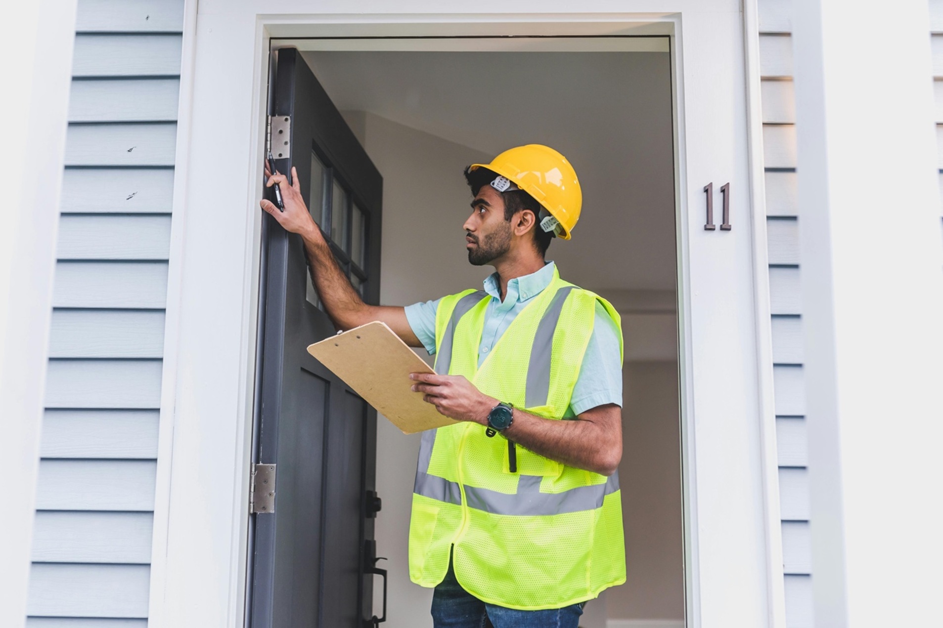 a man in hard hat and neon vest holding a clipboard as he checks the hinges on a front door